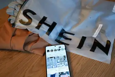 A smartphone showing Shein’s online store lies next to branded packaging and clothing on a wooden table.